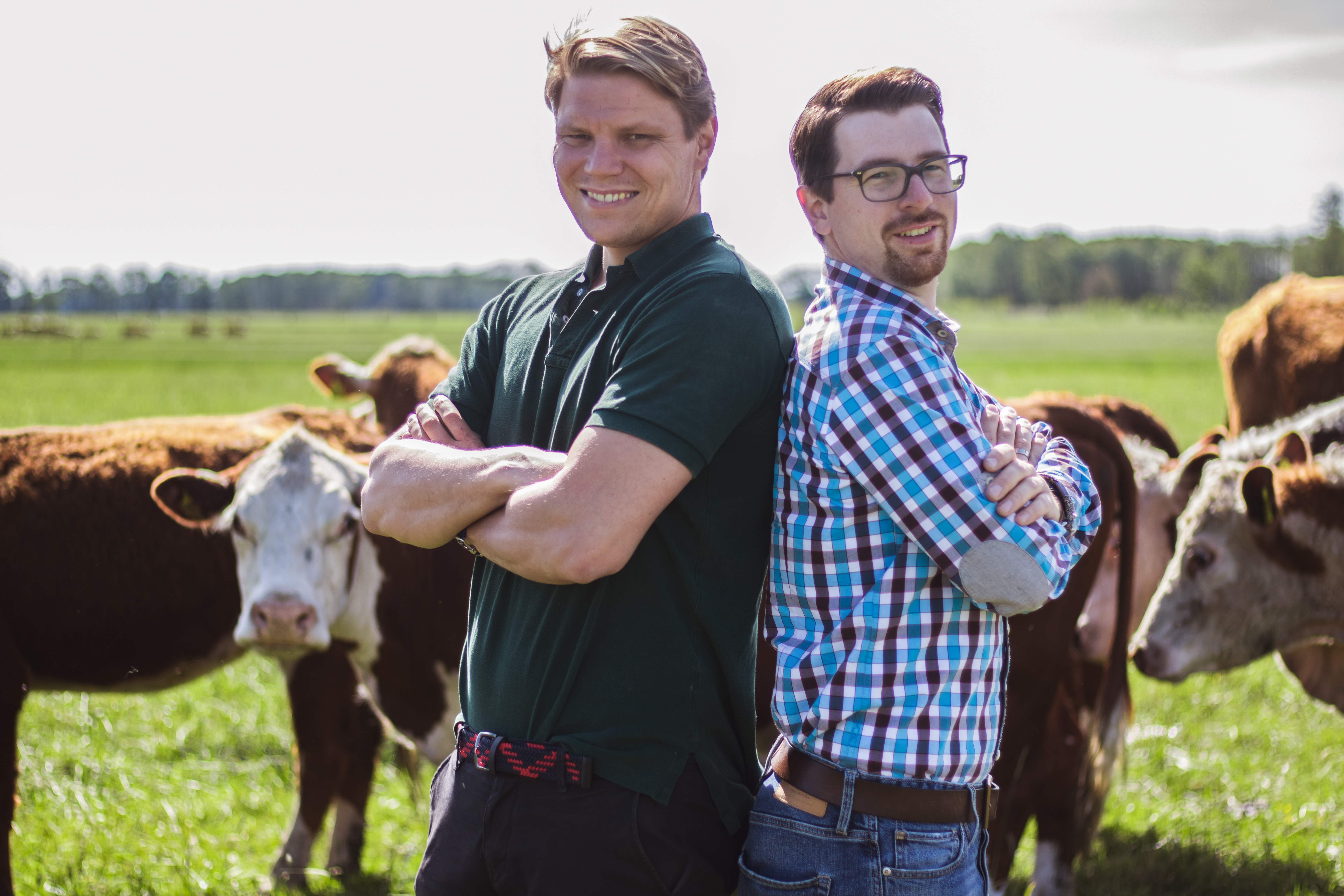 Lorenz Jandke und Jan Heilmann stehen Rücken an Rücken mit den Armen verschränkt vor einer Rinderherde und lächeln in die Kamera.